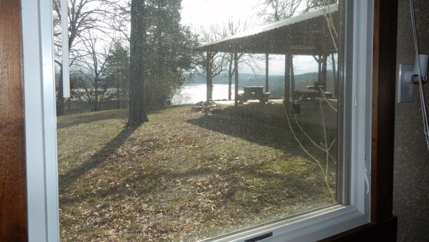 A view through a window of a covered outdoor area with a picnic table and benches, a grassy area, and trees. - Spacious Norfork Lake View Bunkhouse, Resort Amenities, Family/Fishermen/Pets (Gamaliel)