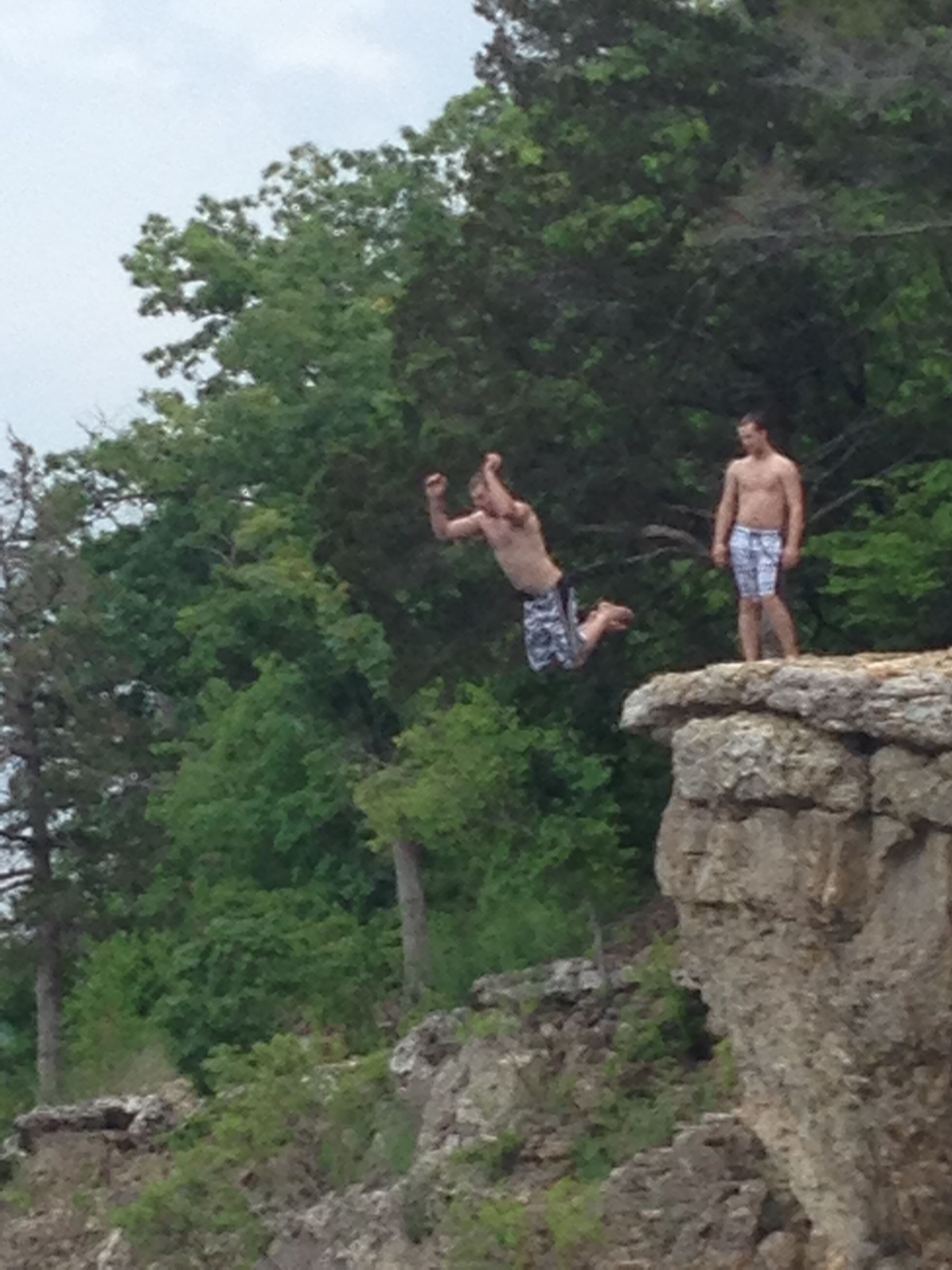 A man jumping off a rocky cliff into a forested area.