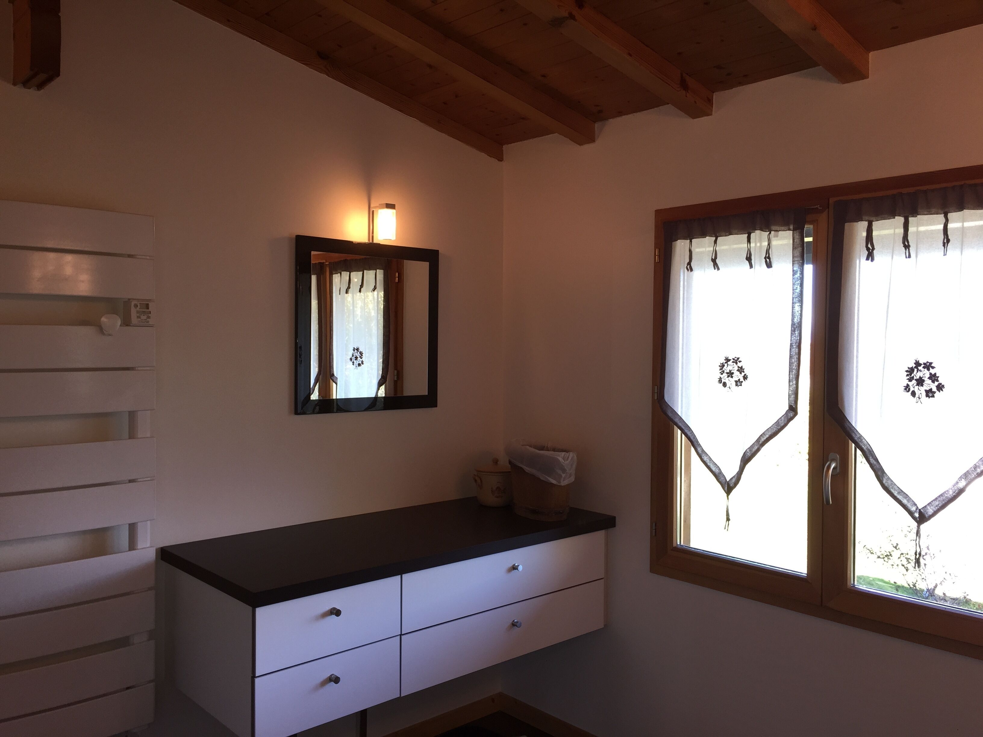 Una habitación con techo de madera, un aparador blanco con cajones, un espejo y una ventana con cortinas decorativas.