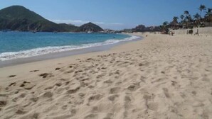 Une plage de sable bordée d’une eau d’un bleu limpide, une montagne en arrière-plan et quelques bâtiments le long du rivage.