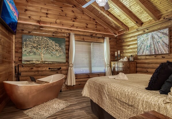 A log cabin bedroom with a large bed, a bathtub, and a painting of a tree.