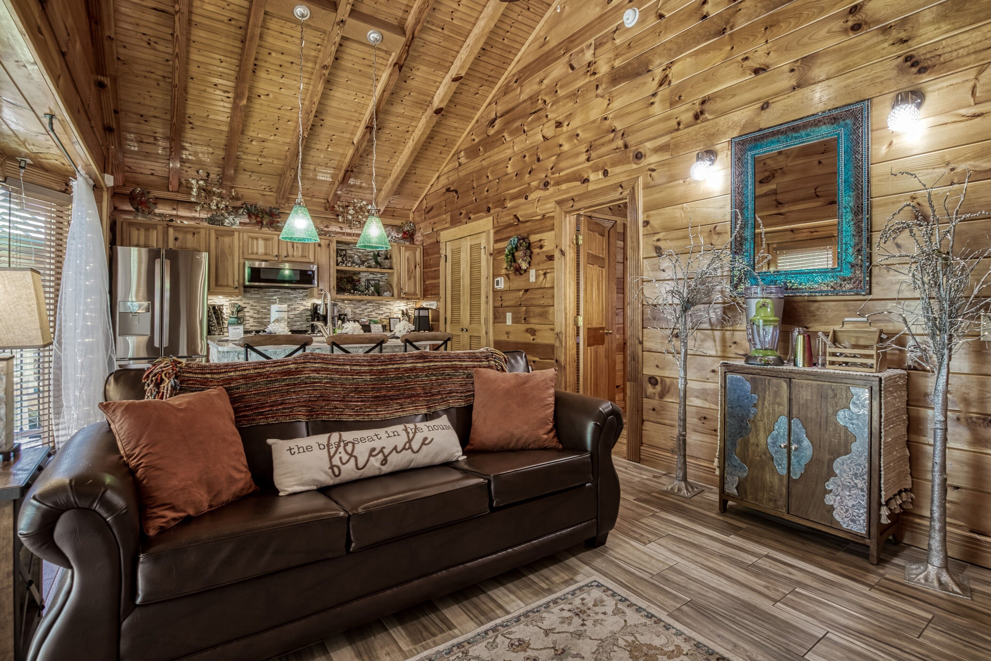 A cozy cabin interior with a wooden ceiling, a leather sofa, a dining table, and a kitchen area.