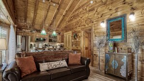 A cozy cabin interior with a wooden ceiling, a leather sofa, a dining table, and a kitchen area.