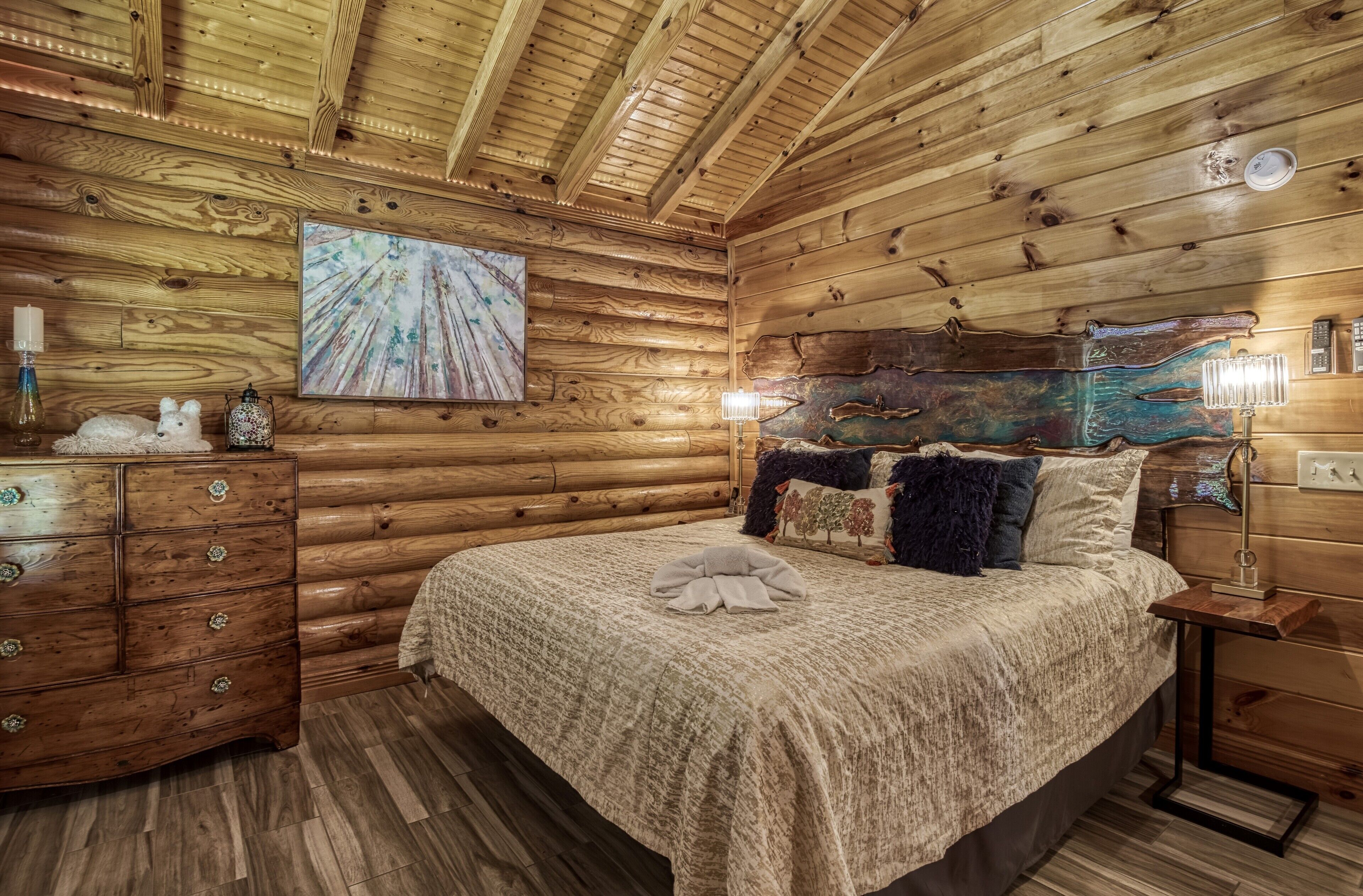 A cozy log cabin bedroom with a wooden ceiling, a bed with a blanket, a dresser with drawers, and a wall-mounted lamp.