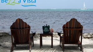 Två trästolar står vända mot en sandstrand med utsikt över havet, och en segelbåt syns på avstånd. En grön väska med en flaska står på ett litet bord mellan stolarna.