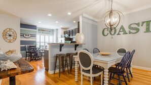 A dining area with a white table, dark chairs, and a chandelier. A kitchen is visible in the background with a 'COAST' sign on the wall.