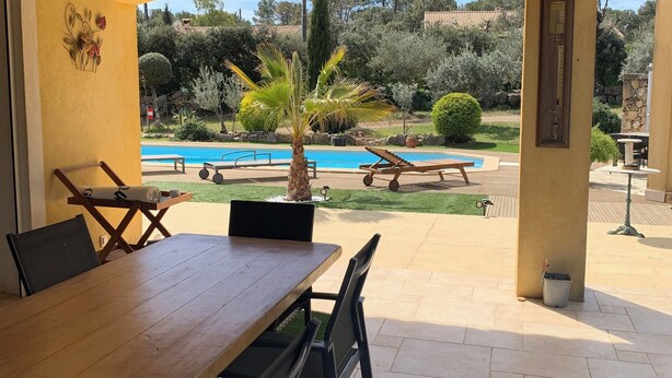 An outdoor patio with a wooden table and chairs, a pool, and a view of the surrounding garden.