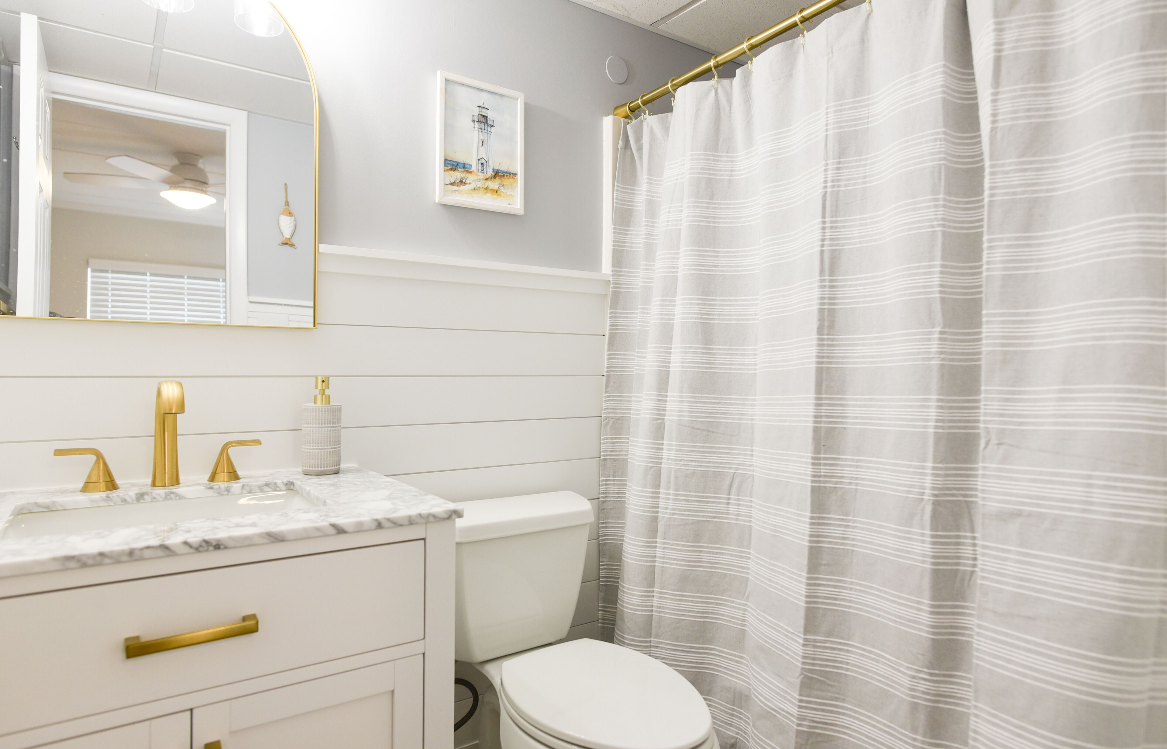 Une salle de bain avec des toilettes blanches, un lavabo avec un robinet doré, un comptoir en marbre blanc, un porte-serviettes doré, un rideau de douche blanc et un tableau encadré au mur.