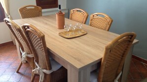 Une table à manger en bois, avec quatre chaises en osier, une bouteille et deux verres à vin posés sur un set de table.