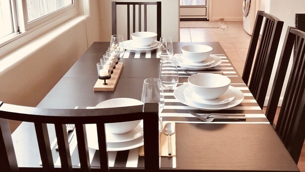 A dining area with a long table set for six, flanked by chairs, with a view of a laundry room through an archway.