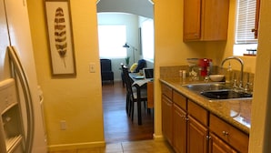 A kitchen with wooden cabinets, a granite countertop, and a sink. There is a framed picture on the wall and a plant on the dining table.