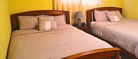 Una habitación con dos camas de madera, una mesita de noche con lámpara y un ventanal con cortinas.