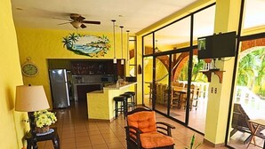 Una sala de estar con un sofá, sillones, una mesa de centro y un televisor. Se ve un área de cocina al fondo, y un comedor a través de amplias ventanas.