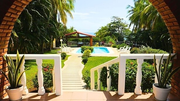 Vista a través de un arco de ladrillo hacia una zona de piscina con un área de estar cubierta y un jardín cuidado.