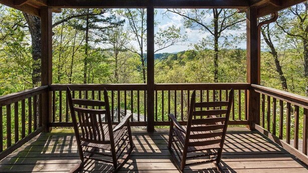 Um deck de madeira com duas cadeiras e uma mesa, com vista para uma paisagem arborizada.