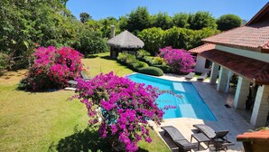 Una piscina rodeada de exuberante follaje y flores vibrantes, con sillones de descanso y una estructura de techo de paja cerca.