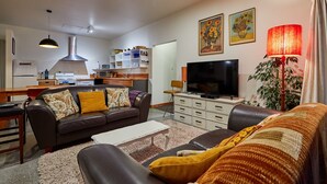 A modern living room with a sofa, coffee table, TV, and a dining area with a kitchen in the background. - Three bedroom cottage with stunning panoramic views of the sea and mountains. (Kaikoura)