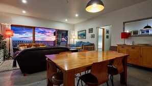A modern living room with a wooden dining table, black leather sofa, and a view of the sunset through large windows. - Three bedroom cottage with stunning panoramic views of the sea and mountains. (Kaikoura)