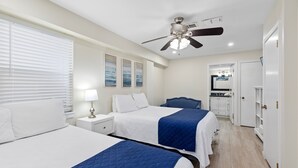 A hotel room with two beds, a ceiling fan, a nightstand with a lamp, a desk, and a view of the kitchen and bathroom through an open door.