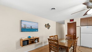 A dining area with a wooden table and chairs, a television mounted on the wall, and a refrigerator in the background.