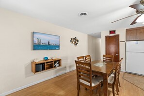 A dining area with a wooden table and chairs, a television mounted on the wall, and a refrigerator in the background. - Multi-Family Beach Retreat • Private Heated Pool • 3 Min Walk to Beach (South Padre Island)