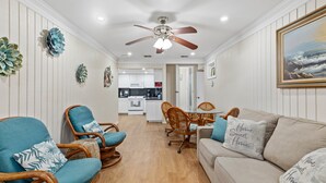 A spacious living room with a beige sofa, teal armchairs, and a wooden dining table. A ceiling fan and recessed lighting are visible.