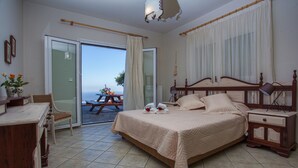 Un dormitorio con una cama grande, una mesita de noche de madera, una mesilla auxiliar, una silla de mimbre, un escritorio y vistas al mar a través de un ventanal corredera.