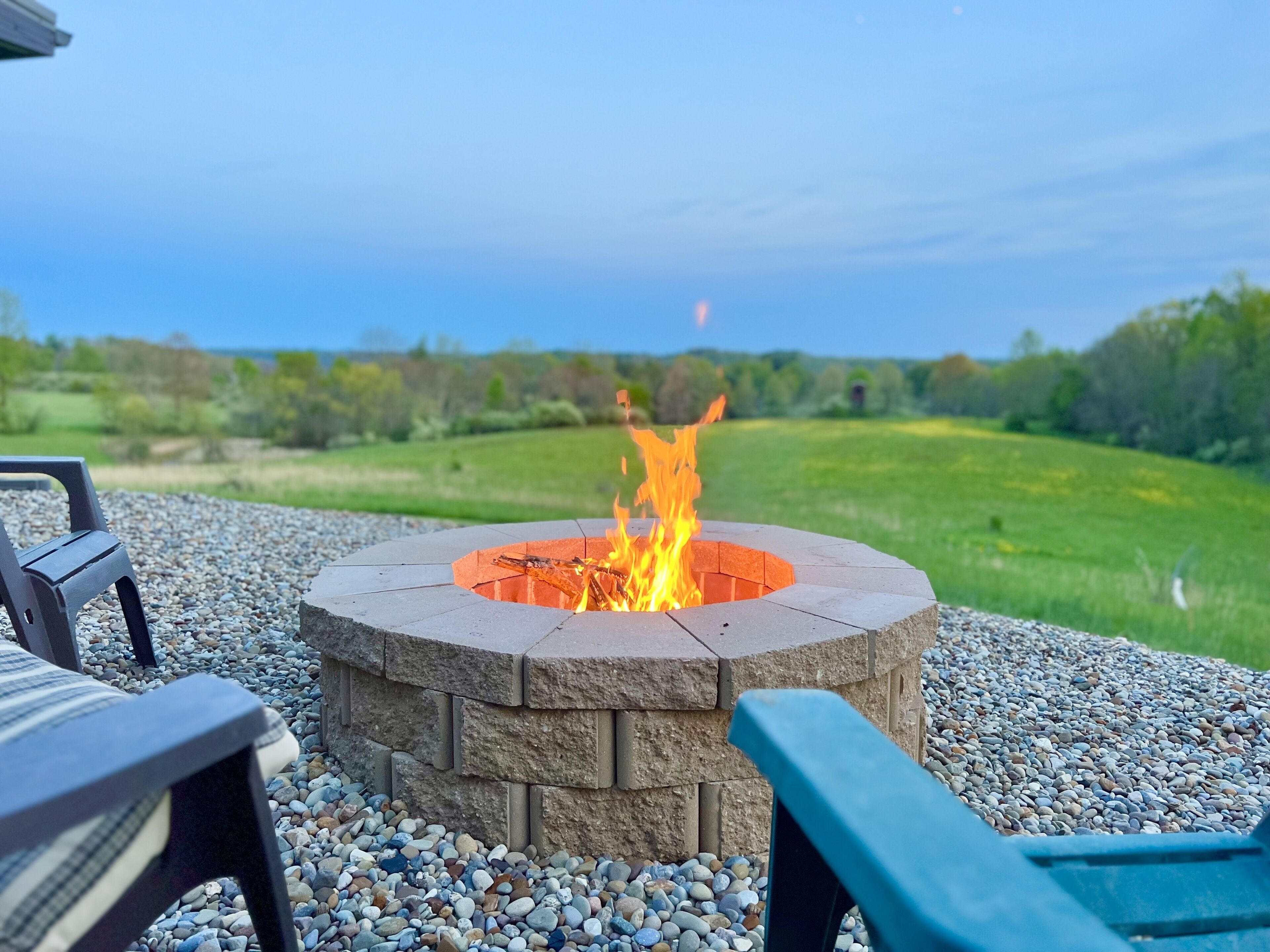 A stone fire pit with a fire burning, surrounded by pebbles and two chairs.