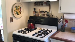 A kitchen with a gas stove, oven, and white cabinets. There is a red kettle on the stove and a plate with fruit on the wall. - Peaceful Escape, Tropical Gardens, Easy Walk to Beach & Eats  (Capitola)