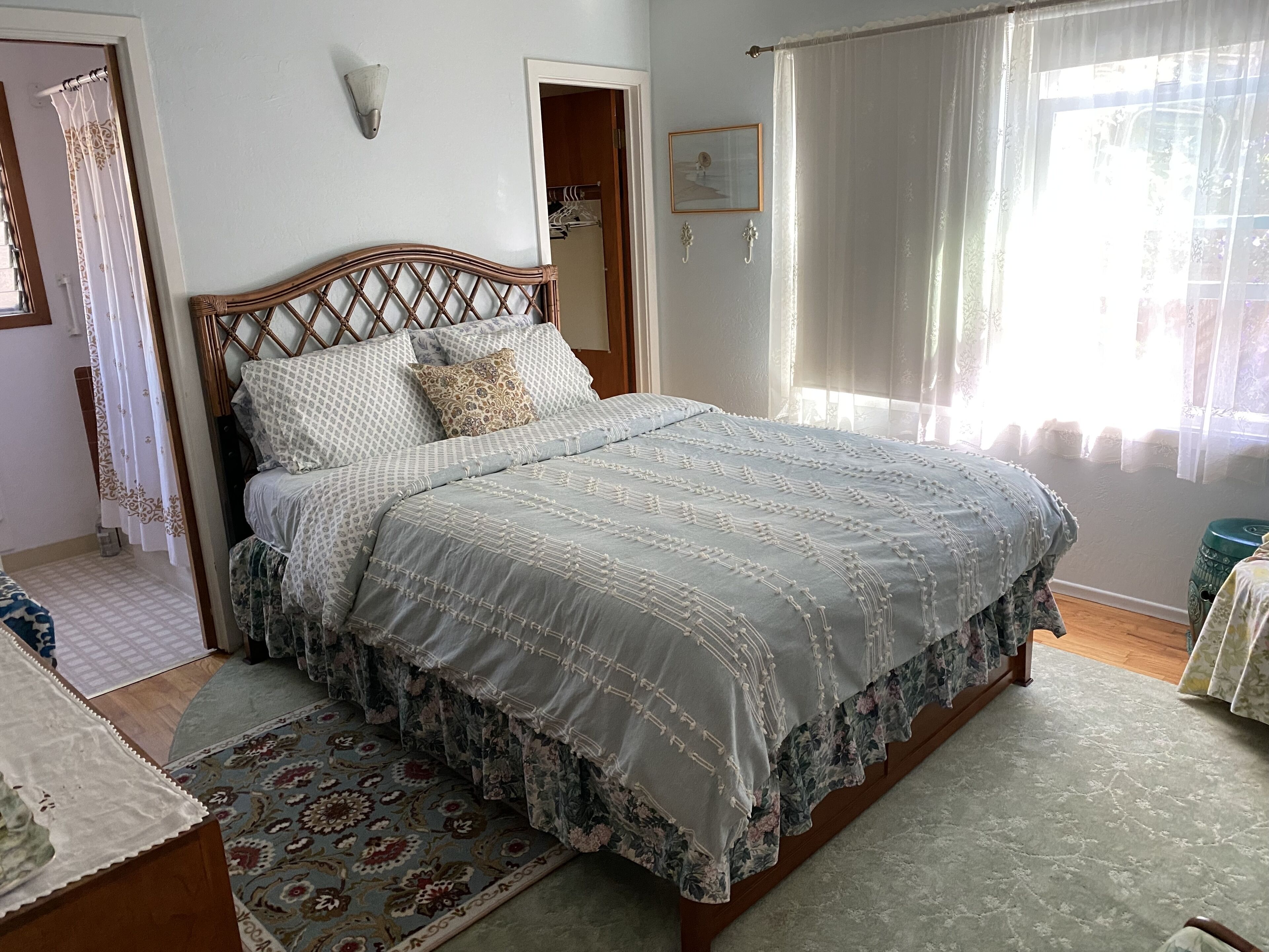 A neatly made bed with a patterned bedspread and pillows, a wooden headboard, a small rug, and a window with sheer curtains.