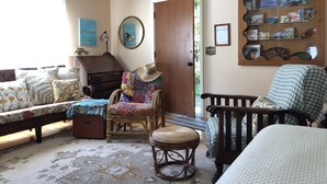A cozy room with a bed, a wooden bench, a wicker chair, a small table, and a bookshelf filled with books and decorative items. - Peaceful Escape, Tropical Gardens, Easy Walk to Beach & Eats  (Capitola)
