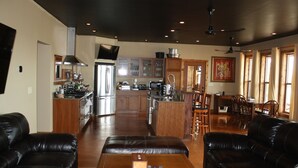 Amplio salón con una gran mesa de centro de madera, sofás de cuero negro y una zona de cocina con armarios de madera y electrodomésticos de acero inoxidable.