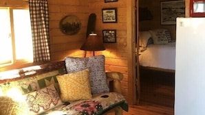 Interior acolhedor de cabana com teto de madeira, sofá com almofadas estampadas, luminária, quadros nas paredes e uma geladeira.