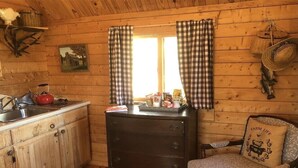 Interior rústico de cabana, com decoração em madeira, área de cozinha com pia e chaleira, poltrona com almofada e janela com cortinas xadrez.
