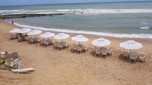 Uma praia com fileiras de guarda-sóis e cadeiras brancas, um molhe rochoso e o oceano ao fundo.