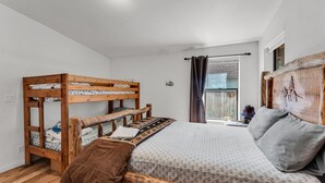 Un dormitorio con literas, una cama grande con ropa de cama estampada, una ventana con cortinas y un cabecero de madera.