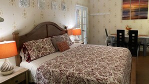 A bedroom with a large bed, two bedside lamps, a wooden headboard, a dining area with a table and chairs, and a decorative wall with framed artwork.