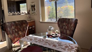 A dining area with a round glass table, floral patterned chairs, a decorative bow on the window, and a view of autumn trees.
