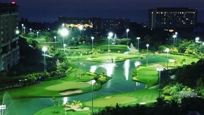 Un campo de golf al anochecer con luces iluminadas, obstáculos acuáticos y edificios circundantes.