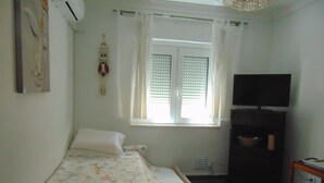 A bedroom with a bed, a television, a window with blinds, and a wall-mounted air conditioner. - Rio ... Beaches ... Lisbon, Ideal for families with children and pets. (Almada)