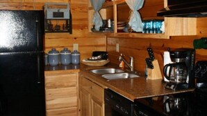 A compact kitchen with wooden cabinets, a black refrigerator, a coffee maker, and a dishwasher. - Morningstar Cabin Caddo River. GREAT VALUE!!!!!!!!!!! River access!! (Glenwood)