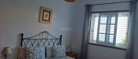 Een slaapkamer met een bed, nachtlampjes, een hanglamp, een raam met gordijnen en een ingelijst schilderij aan de muur.