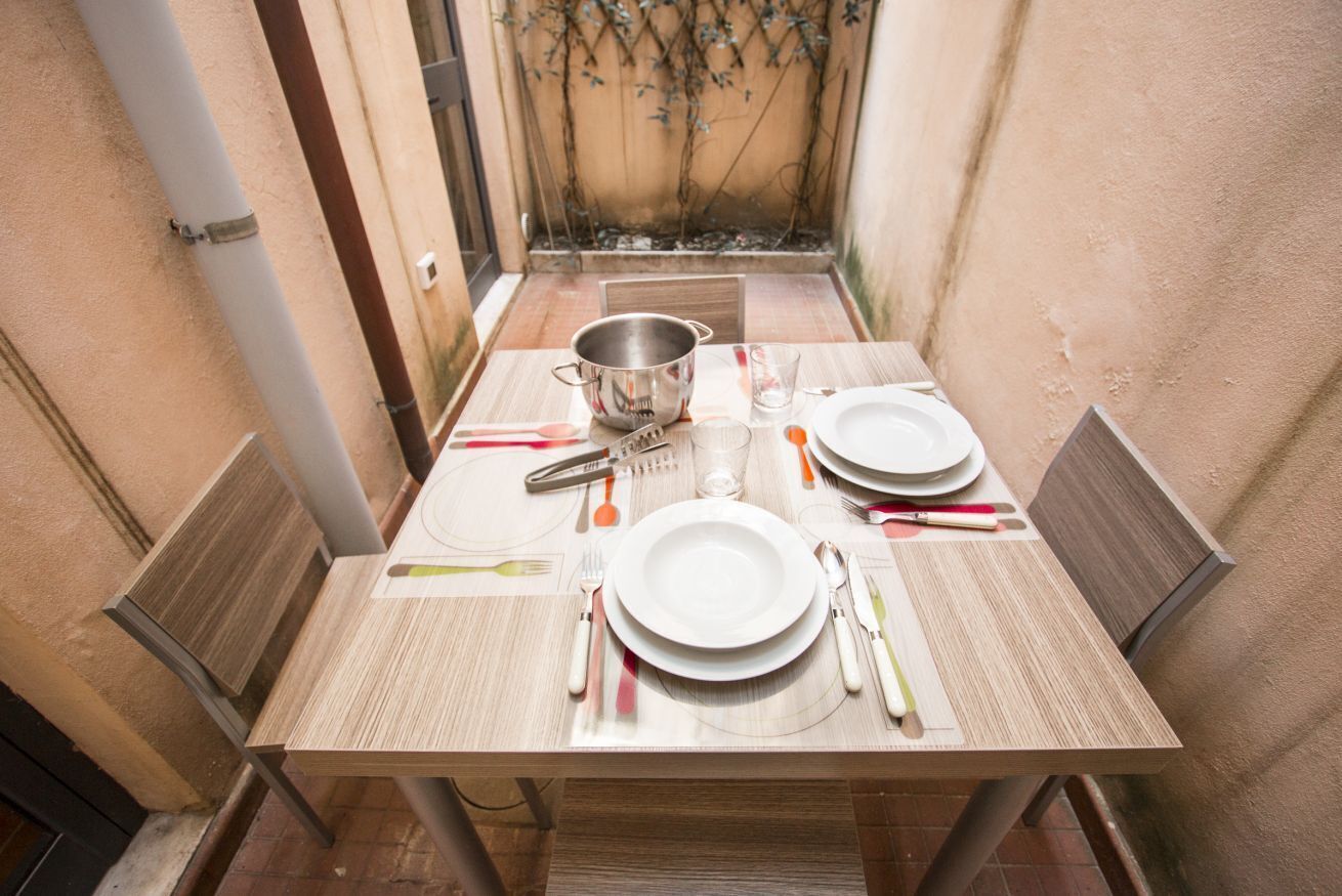 A dining table set with plates, cutlery, and a pot, surrounded by chairs in a small, enclosed space with a tiled floor and a wall with climbing plants.