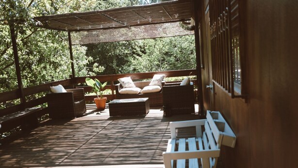 Terraza de madera con mobiliario exterior, que incluye un banco y dos sillones con cojines, rodeada de plantas en macetas y un pérgola.