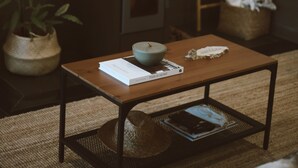 Una habitación pequeña y acogedora con una mesa de madera, un cuenco y un libro. Hay una planta, una cesta tejida y una escalera con una toalla colgada.