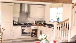Cocina con armarios blancos, extractor de acero inoxidable, microondas y estufa. Hay un comedor con mesa y sillas, y una escalera que sube a otro nivel.