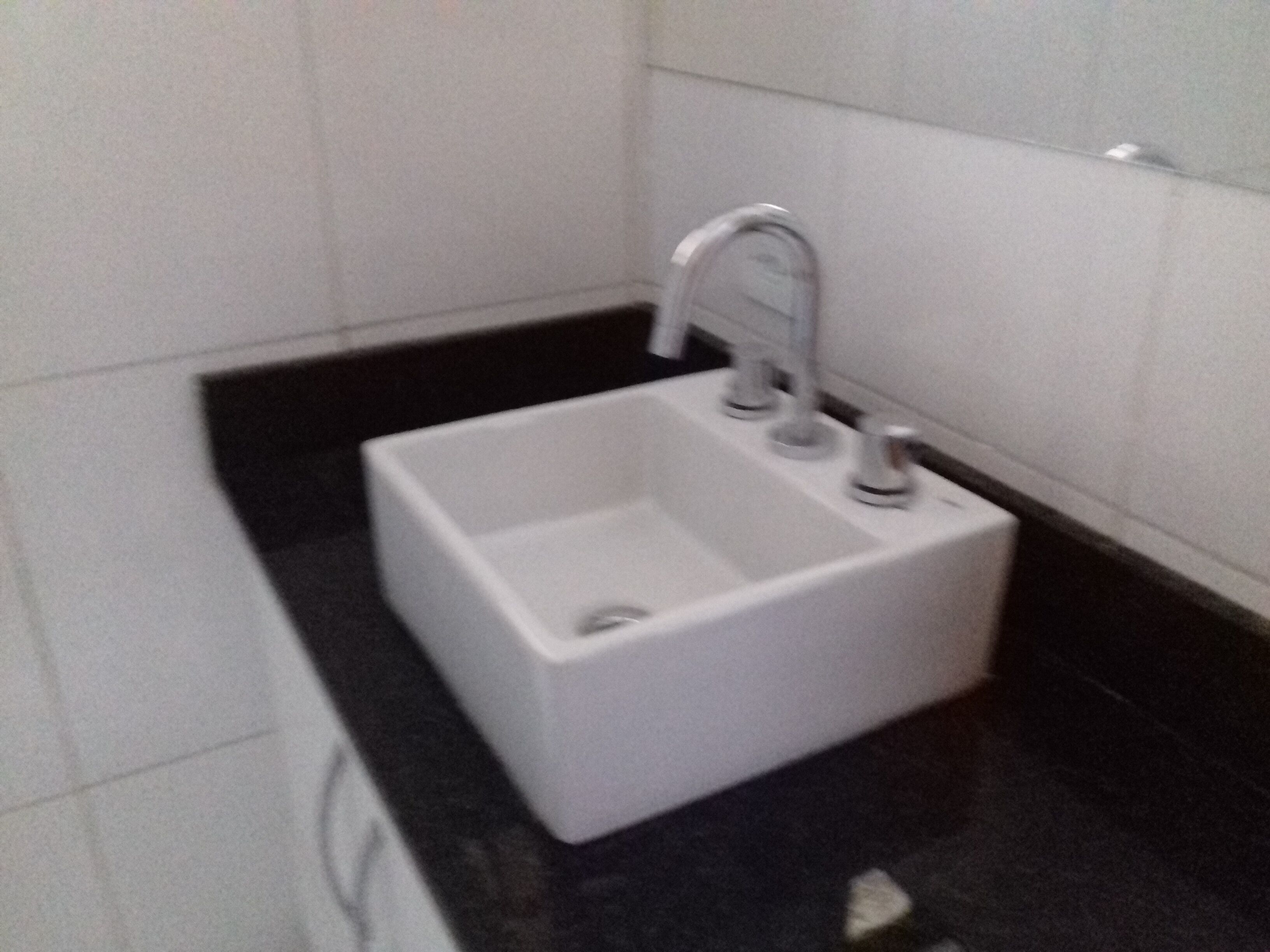 A small, white, wall-mounted sink with a single faucet in a bathroom with black flooring and white tiles.