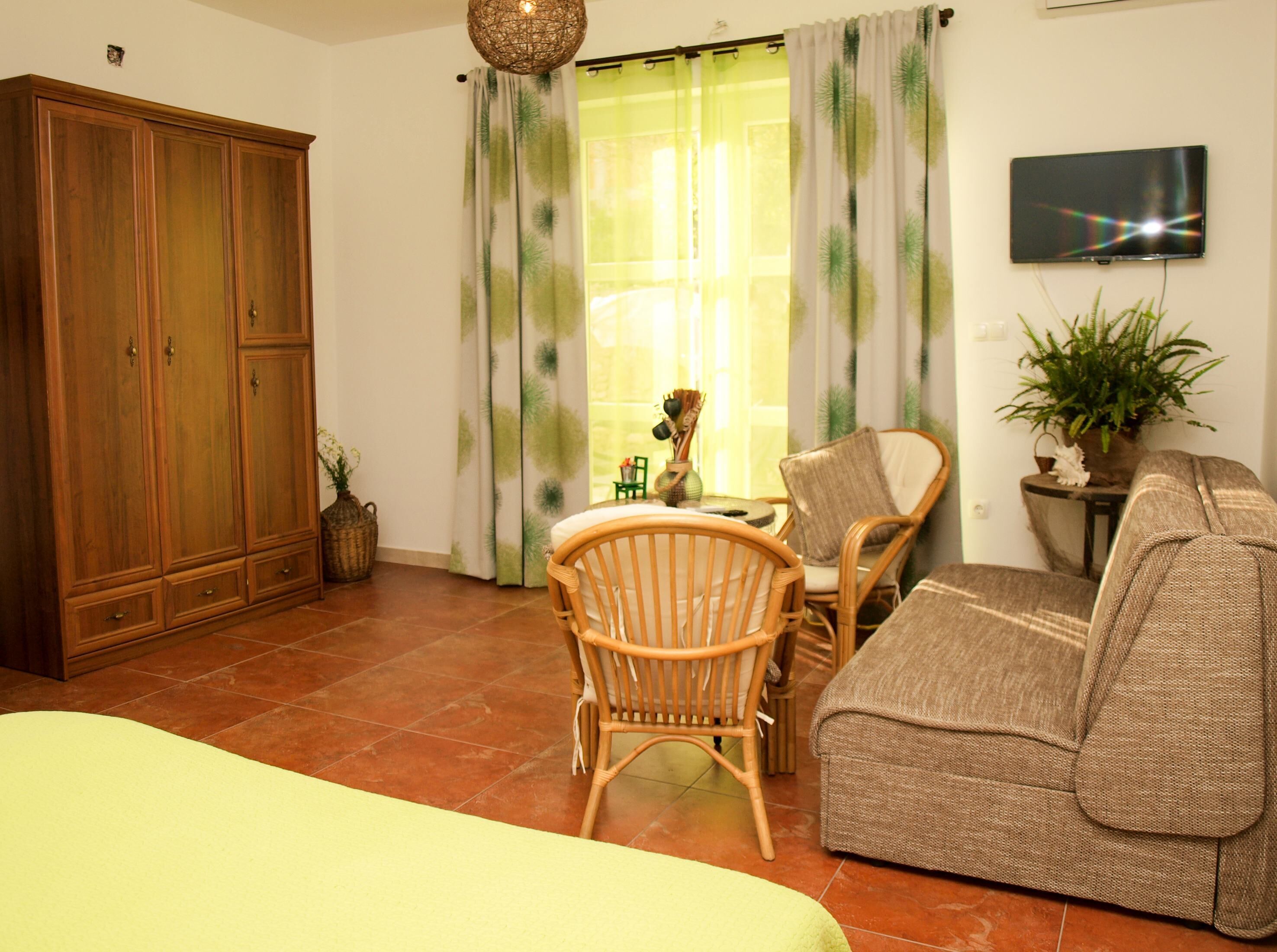 A living room with a wooden wardrobe, a wicker chair, a sofa, a television on the wall, and a window with curtains.