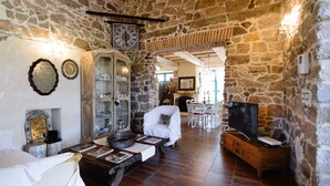Una habitación con paredes de piedra, suelo de madera, un sillón blanco, un armario de madera y un televisor sobre un mueble de madera.