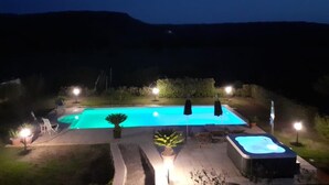 Una piscina con jacuzzi, rodeada de sillas de descanso y plantas en macetas, iluminada con luces nocturnas.